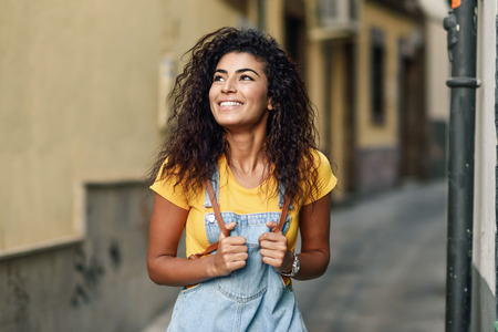 Young Arab tourist woman with black curly hairstyle outdoors. North African traveler girl in casual clothes in the street. Happy female wearing yellow t-shirt and denim dress in urban background.の写真素材
