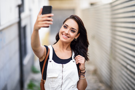 Young woman taking selfie photograph with her smart phone outdoors. Girl wearing denim dress in urban background. Technology concept.の写真素材