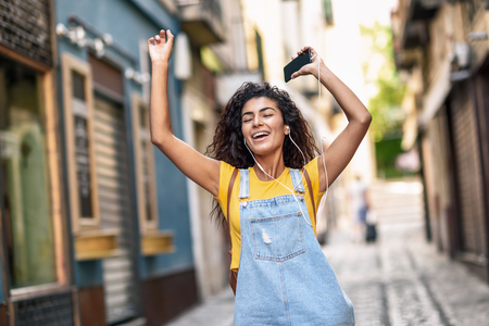 Funny black woman listening to music with earphones outdoors. Arab girl in casual clothes with curly hairstyle in urban background.の写真素材