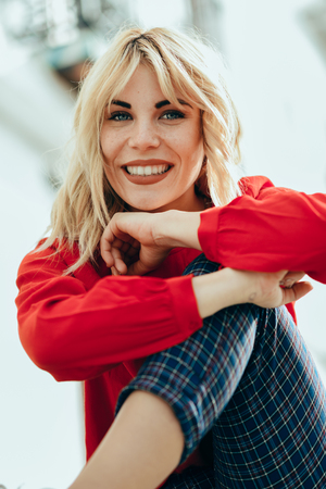 Happy young blond woman sitting on urban background. Smiling blonde girl with red shirt enjoying life outdoors.の写真素材