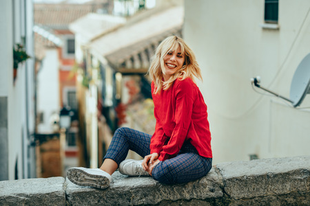 Happy young blond woman sitting on urban background moving her hair. Smiling blonde girl with red shirt enjoying life outdoors.の写真素材