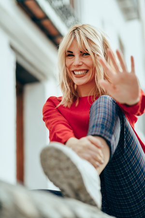 Happy young blond woman sitting on urban background. putting her hand near the camera.の写真素材