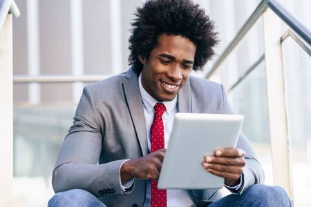 Black Businessman using a digital tablet sitting near an office building.の写真素材