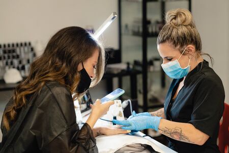 Masked beautician, painting her clients nails while she records a video with her smartphone.の写真素材