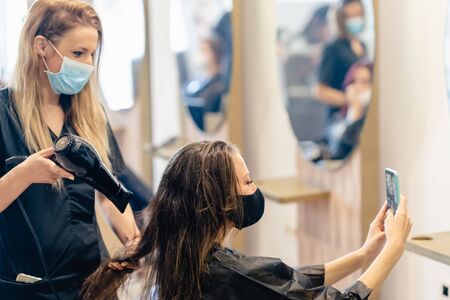 Hairdresser drying her clients hair with a hairdryer wearing protective masks in a beauty centre.の写真素材