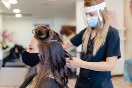 Hairdresser, protected by a mask, combing her clients hair with a hair iron in a salon.の写真素材