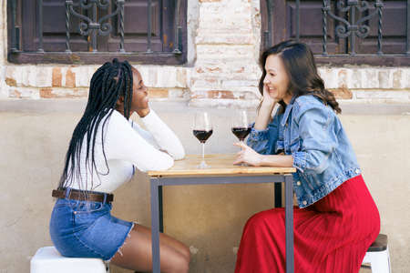 Two multiethnic friends talking sitting at a table outside a bar while drinking a glass of red wine.の写真素材