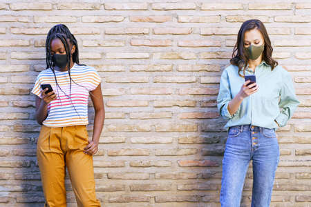 Multiethnic young women wearing masks using smartphone separated to respect social distance.