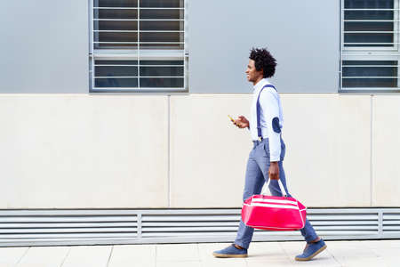 Black man with afro hairstyle carrying a sports bag and smartphone outdoors.の写真素材