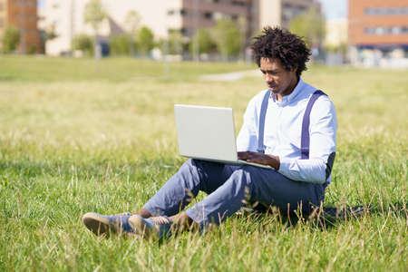 Black man with afro hair using his laptop sitting on skateboard on the grass of an urban park.の写真素材