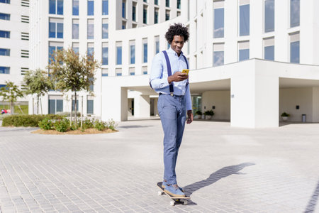 Black businessman on a skateboard looking at his smartphone outdoors.の写真素材