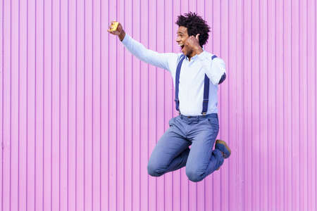 Black man taking a selfie with his smartphone while jumping outdoors.の写真素材