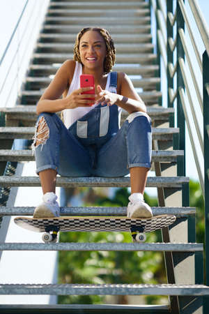 Black girl with coloured braids, sitting with a smartphone and a skateboard.の写真素材