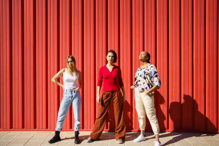Stylish young diverse ladies standing against red fence on streetの写真素材