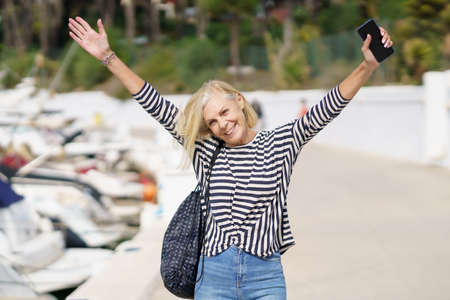 Woman in her 60s strolling along a seaside spot near the beach, raising her arms in joy.