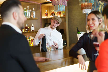 Group of well dressed friends drinking alcohol beverages and enjoying meeting while standing at wooden counter in bar with bartenderの写真素材