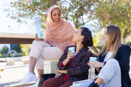 Happy young multiethnic female friends, in casual clothes, smiling while resting in outdoor bench and drinking coffee from take-away cupsの写真素材
