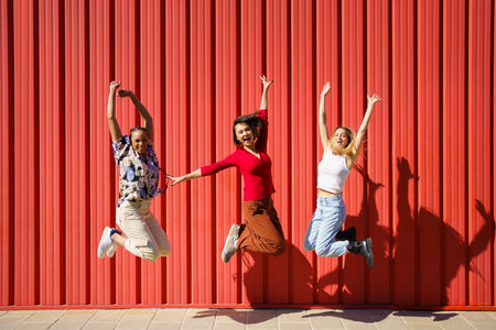 Full body of joyful multiethnic female friends looking at camera and jumping near red wall while having fun on sunny dayの写真素材