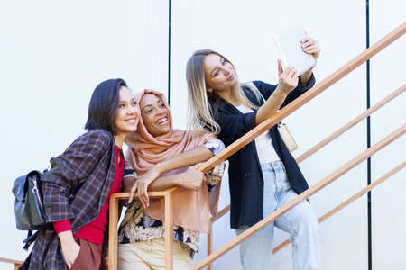 Low angle of delighted young multiracial female students in trendy outfit gesturing and smiling while taking selfie on tablet standing on stairs on city streetの写真素材