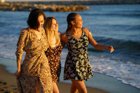 Joyful young multiracial female friends in sundresses walking together on sandy coast near waving sea while enjoying sunny summer dayの写真素材