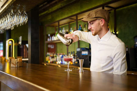 Bearded man in white shirt and cap filling glass with cocktail from shaker while standing behind...