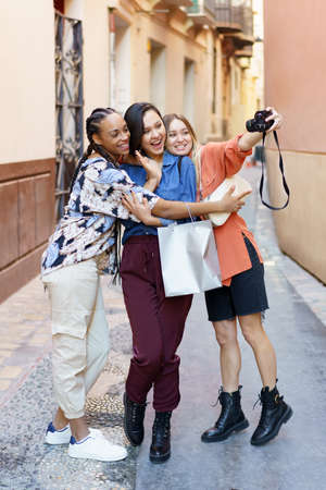 Full length of glad multiracial female friends taking selfie on camera while standing on street near building after shoppingの写真素材