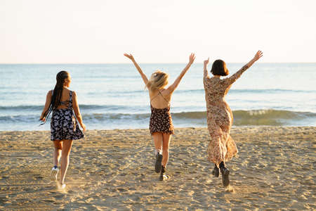 Full body back view of unrecognizable female friends in dresses running on sandy beach with raised arms near waving seaの写真素材