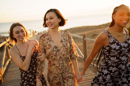 Optimistic multiracial female friends holding hands and walking together on wooden boardwalk while...