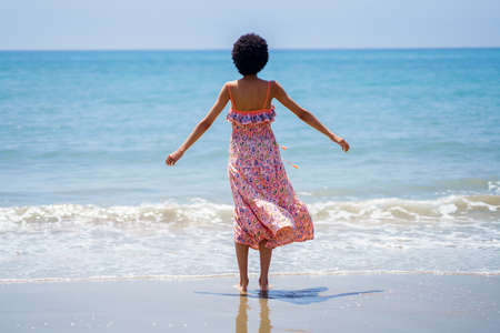 Rear view of black woman with afro hair in summer dress, opening her arms looking out to sea. Girl...