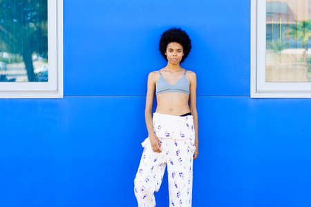 Young black woman with afro hair, fashion model, posing against an urban blue wall, wearing trousers and bra.の写真素材