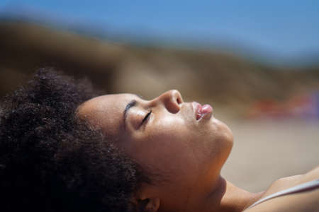 Side view of peaceful black female tourist with Afro dark hair, relaxing on sandy beach with closed eyes, and sunbathing on sunny day during summer holidayの写真素材