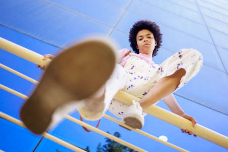 From below full body of African American female in overall and sneakers looking at camera while...