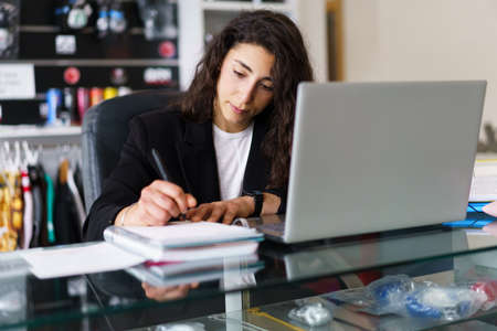 Young sports psychologist sitting at desk with netbook and writing in notepad during therapy session...