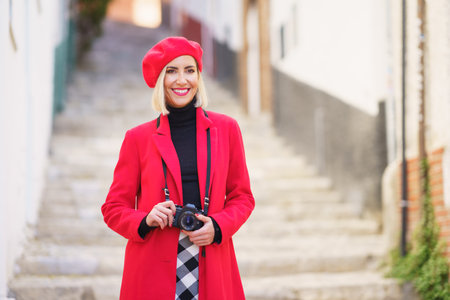 Confident young positive woman, with blond hair in fashionable red coat and beret smiling and looking away while standing on stone stairs with photo camera during trip in old townの写真素材