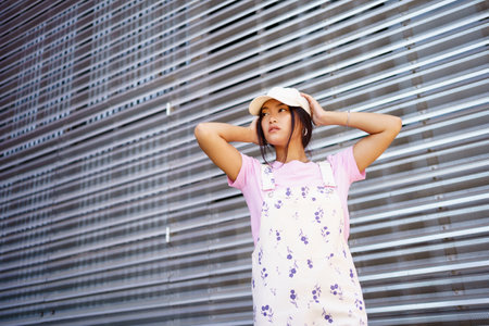 Low angle of glad Asian female in casual clothes, wearing cap, looking away standing near modern...