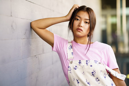Attractive Chinese girl looking at camera with serious expression leaning against the wall of a modern office building. Female wearing casual clothes.の写真素材