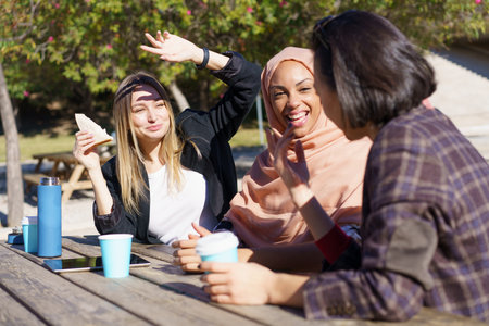 Cheerful young multiracial female coworkers laughing and communicating while having coffee break sitting at wooden table in park on sunny dayの写真素材
