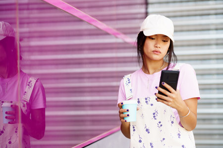 Young Asian female in stylish clothes and cap with takeaway coffee, browsing social media on...