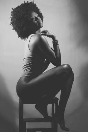 Side view of slim African American female model in bodysuit, with curly hair touching shoulder and looking away while sitting on stool. Black and white photograph.の写真素材
