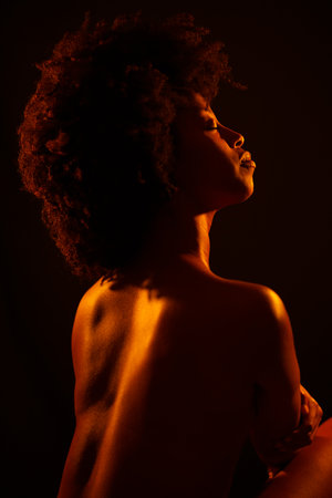 Naked African American woman model with Afro hairstyle closing eyes and enjoying bright neon orange light against black backgroundの写真素材