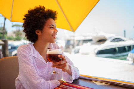 Cheerful young African American female in white blouse with curly hair smiling and looking away, while enjoying beer in street restaurant on sunny summer day on embankmentの写真素材