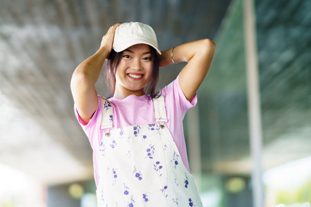 Merry young Asian woman in stylish t shirt and overalls with cap, looking at camera with smile and touching head on blurred background of contemporary building in cityの写真素材
