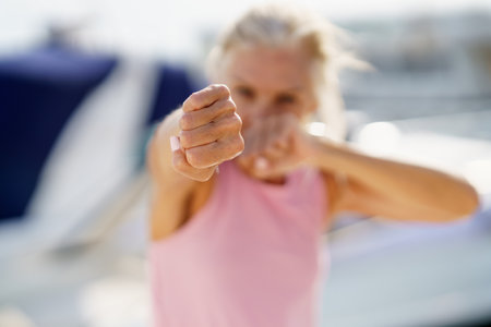 Older woman doing sport in a coastal port. Arm training throwing boxing punches. Mature Female doing...