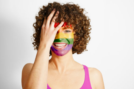 Happy curly haired female with painted in LGBT colors face covering eye looking at camera against white background equality and tolerance conceptの写真素材