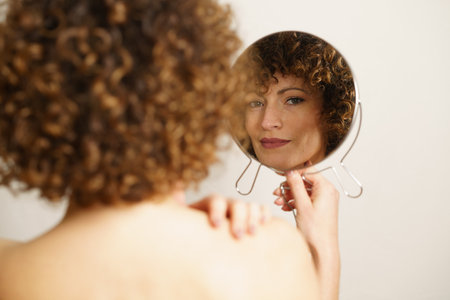 Beautiful young female model, with curly hair and makeup standing with back against camera while holding mirror in hand and looking at camera in reflection near gray backgroundの写真素材