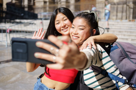 Happy young Asian female travelers, in casual clothes with backpacks smiling and hugging while waving hands and taking selfie on smartphone during summer vacation in Spainの写真素材