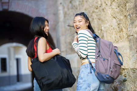 Cheerful Asian female tourists with belongings looking over shoulder at camera with backpack while standing near stone wall of buildingの写真素材