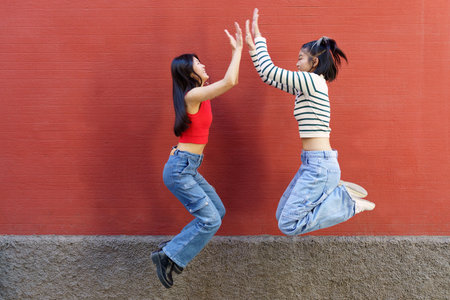 Full body side view of cheerful young Asian female friends jumping up and giving high five to each...