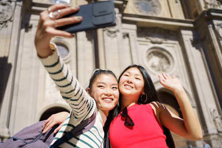 Happy young Asian female travelers, in casual clothes with backpacks smiling and hugging while waving hands and taking selfie on smartphone during summer vacation in Spainの写真素材