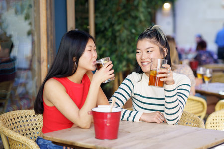 Cheerful young Asian female friends in casual wear sitting at wooden table and chatting while drinking refreshing cold tea during meeting in cafeの写真素材